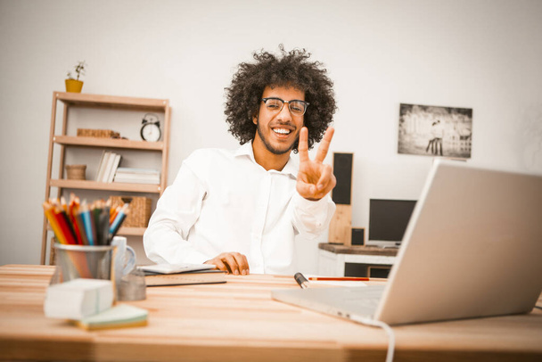 Shaggy creative man shows V or Victory sign or gesture working computer at home interior. Charming student or businessman in eyeglasses smiling at camera. Freelance work concept. Tinted image - Fotoğraf, Görsel