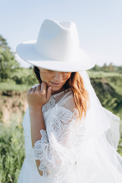 portrait of stylish bride in fedora hat and white dress standing  in the meadow - Фото, зображення