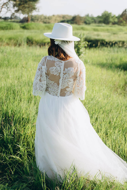 back view of stylish woman wearing fedora hat in white dress standing  in the field  - Fotó, kép