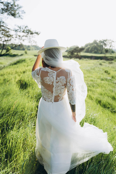 back view of stylish woman wearing fedora hat in white dress standing  in the field  - Foto, afbeelding