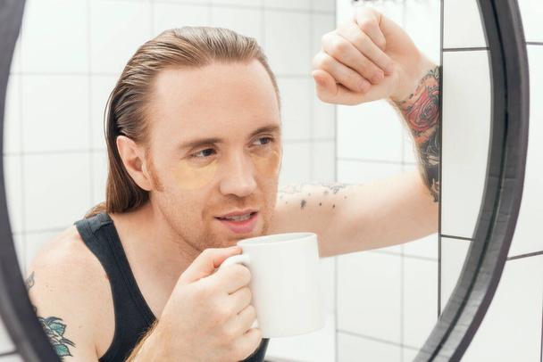 Ένας αξύριστος άντρας με μπαλώματα κάτω από τα μάτια του και καφέ κοιτά τον εαυτό του στον καθρέφτη του μπάνιου.. - Φωτογραφία, εικόνα