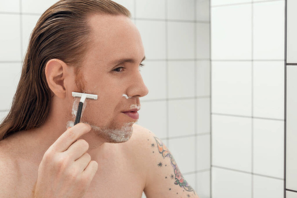 Τατουάζ άντρας με μακριά μαλλιά ξυρίζει το πρόσωπό του μπροστά στον καθρέφτη του μπάνιου..  - Φωτογραφία, εικόνα
