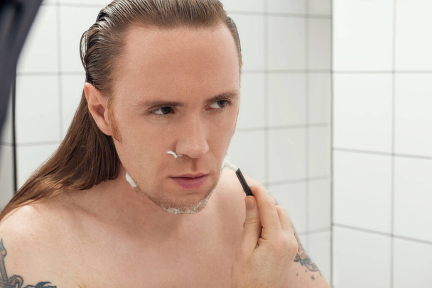 Tattooed man with long hair shaving his face in front of the bathroom mirror.  - Photo, Image
