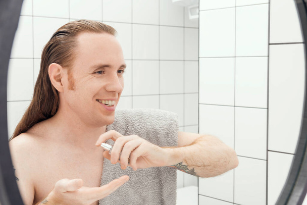 Ein Mann mit Tätowierungen und langen Haarsprays auf der Hand nach dem Rasieren im Badezimmer - Foto, Bild