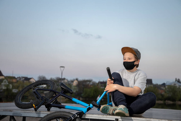 Πορτρέτο ενός εφήβου στο ηλιοβασίλεμα με μια μαύρη μάσκα σε ένα μπλε ποδήλατο. Καραντίνα. Απαγορεύεται η οδήγηση ποδηλάτου. Covid-19. - Φωτογραφία, εικόνα
