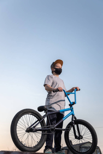 Porträt eines Teenagers im Sonnenuntergang mit schwarzer Maske auf einem blauen Fahrrad. Quarantäne. Es ist verboten, mit dem Fahrrad zu fahren. Covid-19. - Foto, Bild