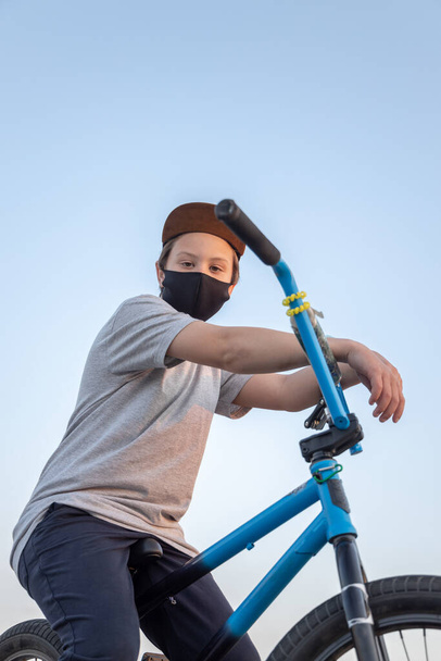 Portret nastolatka w zachodzie słońca w czarnej masce na niebieskim rowerze. Kwarantanna. Zabrania się jazdy na rowerze. Kowid-19. - Zdjęcie, obraz