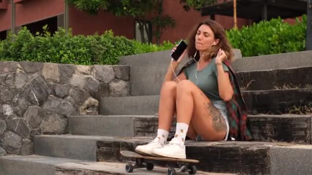 Piękny biały tatuaż pasuje kobieta słuchać muzyki i żuć gumę w jeansowe szorty, koszulka w kratę siedzieć na schodach o zachodzie słońca światło gospodarstwa longboard, deskorolka   - Materiał filmowy, wideo