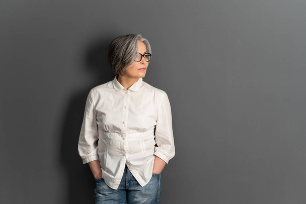 Beautiful mature lady in glasses stands hands in pockets looking at side. Elegant Gray-haired woman in whiite shirt on gray background. Copy space for text at right side - Фото, зображення