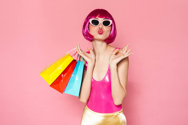 Portrait of an excited young woman wearing colorful bright outfit holding shopping bags isolated over pink background - Zdjęcie, obraz