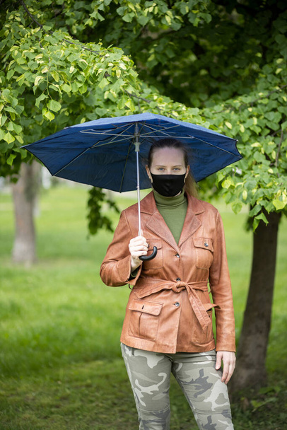 Fekete maszkos nő esernyő alatt az esőben. Egy nő barna kabátban és khaki nadrágban. Zöld fák és fű a háttérben. - Fotó, kép