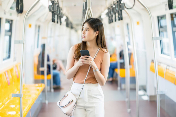 Молода жінка з Азії, пасажирка, користується розумним мобільним телефоном і дивиться на зовнішні поїзди під час подорожі у великому місті, приміських поїздах і транспортних засобах. - Фото, зображення