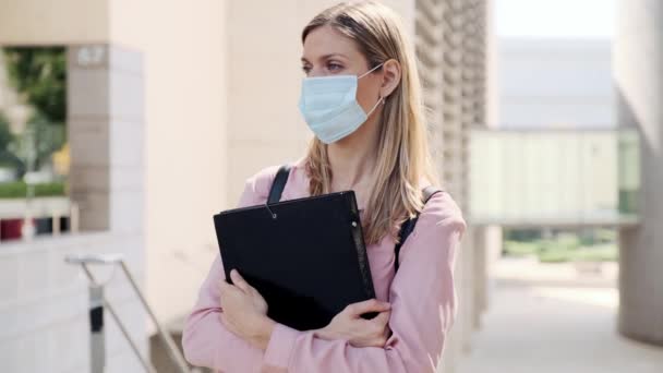 Video of a beautiful blonde woman in face mask with a folder waiting while standing in the street. - Imágenes, Vídeo