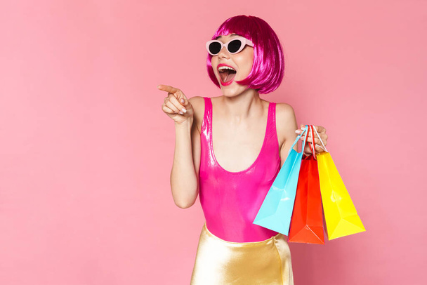 Portrait of an excited young woman wearing colorful bright outfit holding shopping bags isolated over pink background, pointing away - Fotó, kép