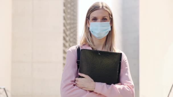 Video of a beautiful blonde woman in face mask with a folder looking at camera while standing in the street. - Filmati, video