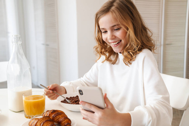 Photo of happy caucasian girl using mobile phone while having breakfast in modern kitchen - Фото, изображение