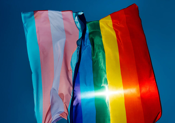 primer plano de una bandera de orgullo gay y una bandera de orgullo transgénero ondeando en el cielo azul, movido por el viento, con el sol en el fondo
 - Foto, imagen