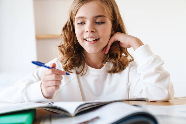 Photo of caucasian happy girl writing in exercise book and smiling while doing homework in living room - Foto, Imagem