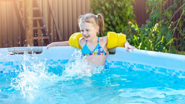 Menina espirra os pés na piscina quadro athome ao ar livre, se divertindo. Ajudas de natação amarelas em criança
 - Foto, Imagem