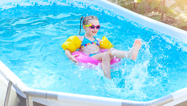 小さな女の子は屋外で楽しんで、自宅でフレームスイミングプールでインフレータブルサークルで飛び散っています。子供の黄色の水泳補助具 - 写真・画像