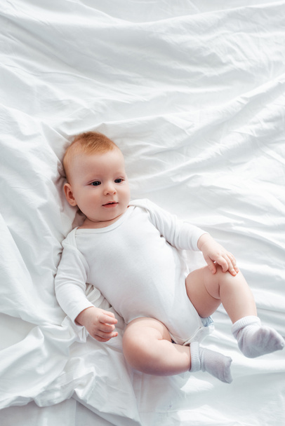 top view of adorable baby boy in romper lying on bedding and looking away - Zdjęcie, obraz