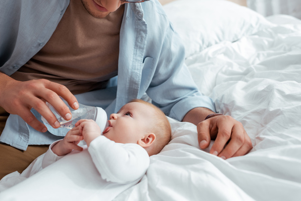 обрезанный вид, как отец кормит очаровательного сына из детской бутылочки на кровати
 - Фото, изображение
