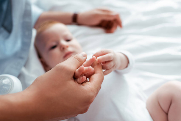 selektywne skupienie uroczego niemowlęcia dotykającego dłoni ojców podczas leżenia w łóżku - Zdjęcie, obraz