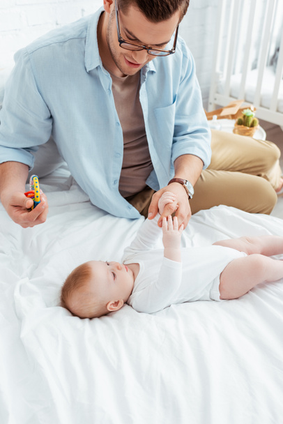 glücklicher junger Vater hält Babyrassel, während er die Hände seines niedlichen kleinen Sohnes berührt - Foto, Bild