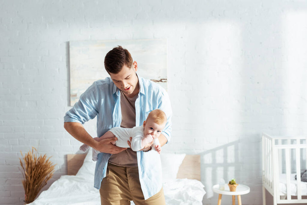 young father holding cute baby boy in romper while standing in bedroom - Photo, Image