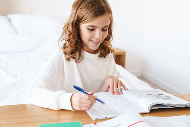 Fénykép kaukázusi boldog lány írásban gyakorló könyv és mosolygós, miközben a házi feladatot a nappaliban - Fotó, kép