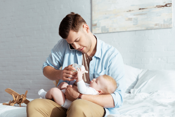 fiatal apa etetés baba fiú cumisüveg, miközben beszél okostelefon - Fotó, kép