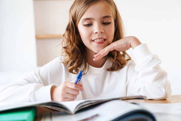 Photo of caucasian happy girl writing in exercise book and smiling while doing homework in living room - Zdjęcie, obraz