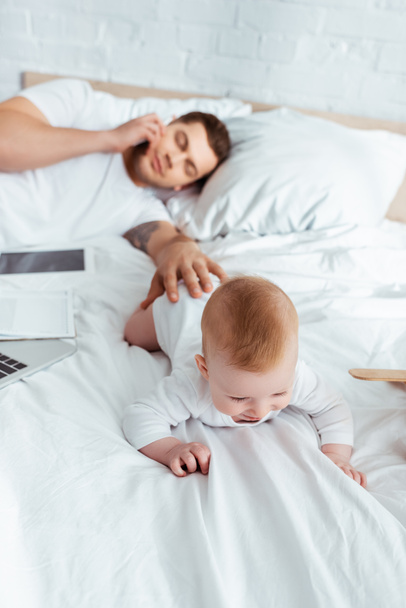 selective focus of man talking on smartphone with closed eyes while touching baby boy crawling on bed - Photo, Image