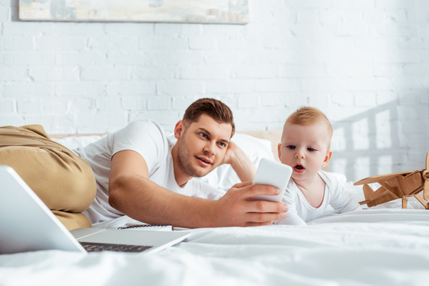 selective focus of young man taking selfie on smartphone with cute little son - Фото, изображение