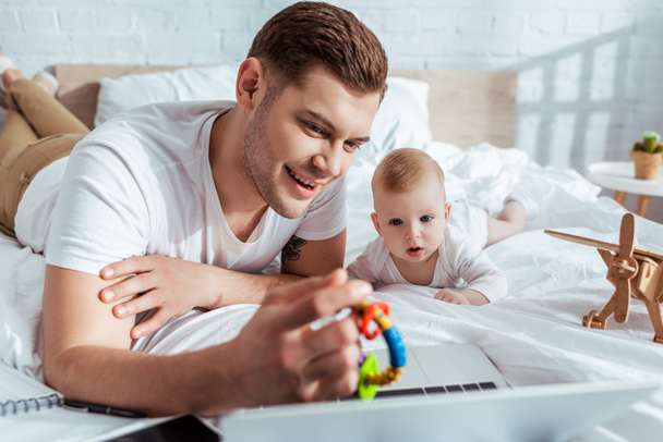 Selektiver Fokus eines lächelnden Vaters, der seinem niedlichen kleinen Sohn Babyrassel zeigt, während er in der Nähe des Laptops im Bett liegt - Foto, Bild