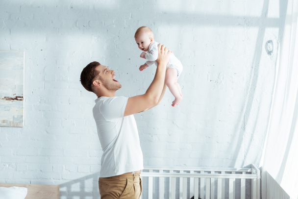 excitado joven padre sosteniendo adorable bebé niño en manos extendidas cerca de cuna
 - Foto, Imagen