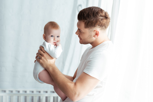 joven padre mirando adorable bebé niño mientras lo sostiene en el dormitorio
 - Foto, imagen