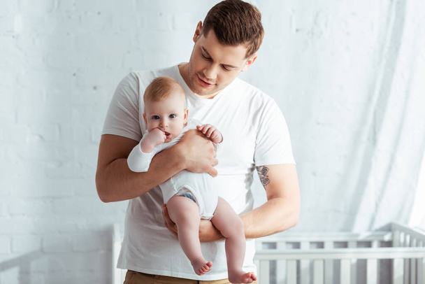 młody ojciec trzyma urocze niemowlę w romper na rękach w sypialni - Zdjęcie, obraz