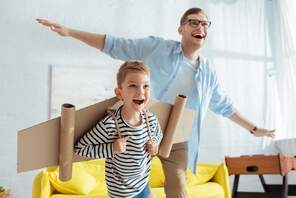 happy boy with carton plane wings, and cheerful father having fun while imitating flying - Fotoğraf, Görsel