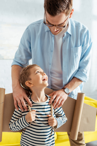 smiling father touching shoulders of happy son with cardboard plane wings on back - Photo, Image