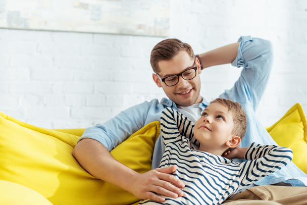happy father and some looking at each other while sitting on sofa at home - Фото, изображение