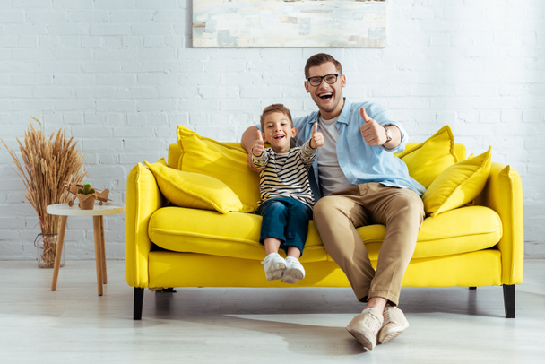 podekscytowany ojciec i syn pokazując kciuki w górę siedząc na żółtej kanapie - Zdjęcie, obraz
