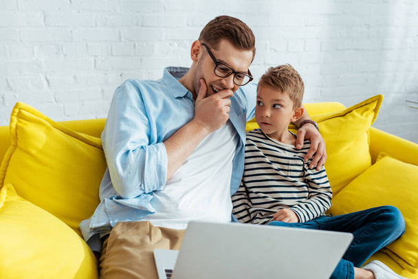 young father laughing and hugging son while sitting on sofa and using laptop - Fotoğraf, Görsel