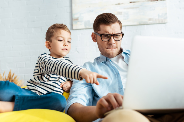 adorable boy pointing with finger at laptop near concentrated father - Fotoğraf, Görsel