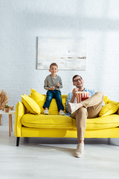 szczęśliwy ojciec i syn siedzi na żółtej kanapie, oglądając telewizję i jedząc popcorn - Zdjęcie, obraz