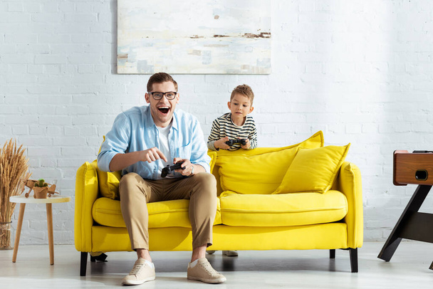 KYIV, UKRAINE - JUNE 9, 2020: excited man playing video game near adorable son - 写真・画像