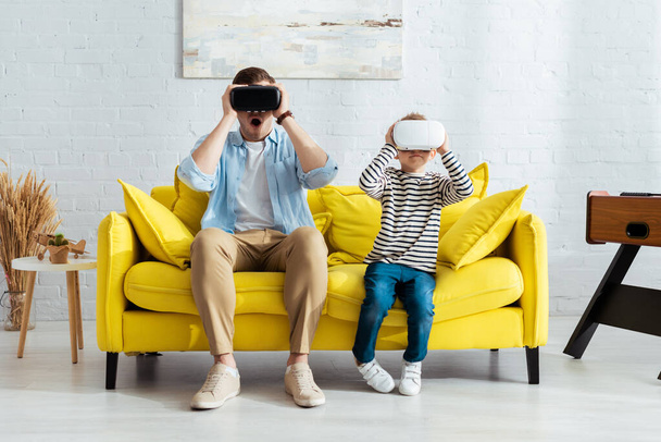 excited father and son sitting on yellow sofa and using vr headsets - Photo, Image