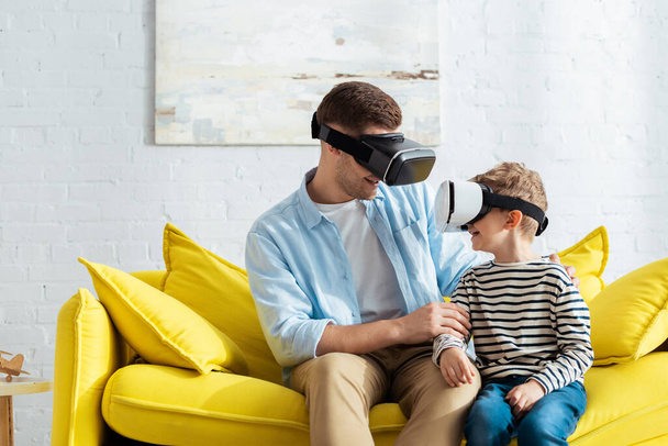 smiling father and son looking at each other while sitting on sofa in vr headsets - Photo, Image