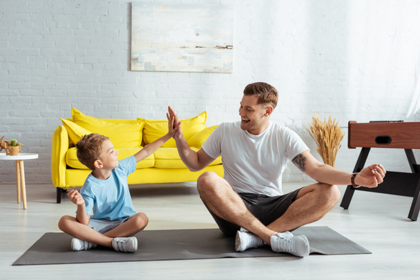 cheerful father and son giving high five while sitting on fitness mat in lotus poses - Foto, afbeelding