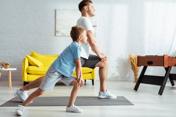 side view of young father and cute son doing lunges while exercising at home - Zdjęcie, obraz